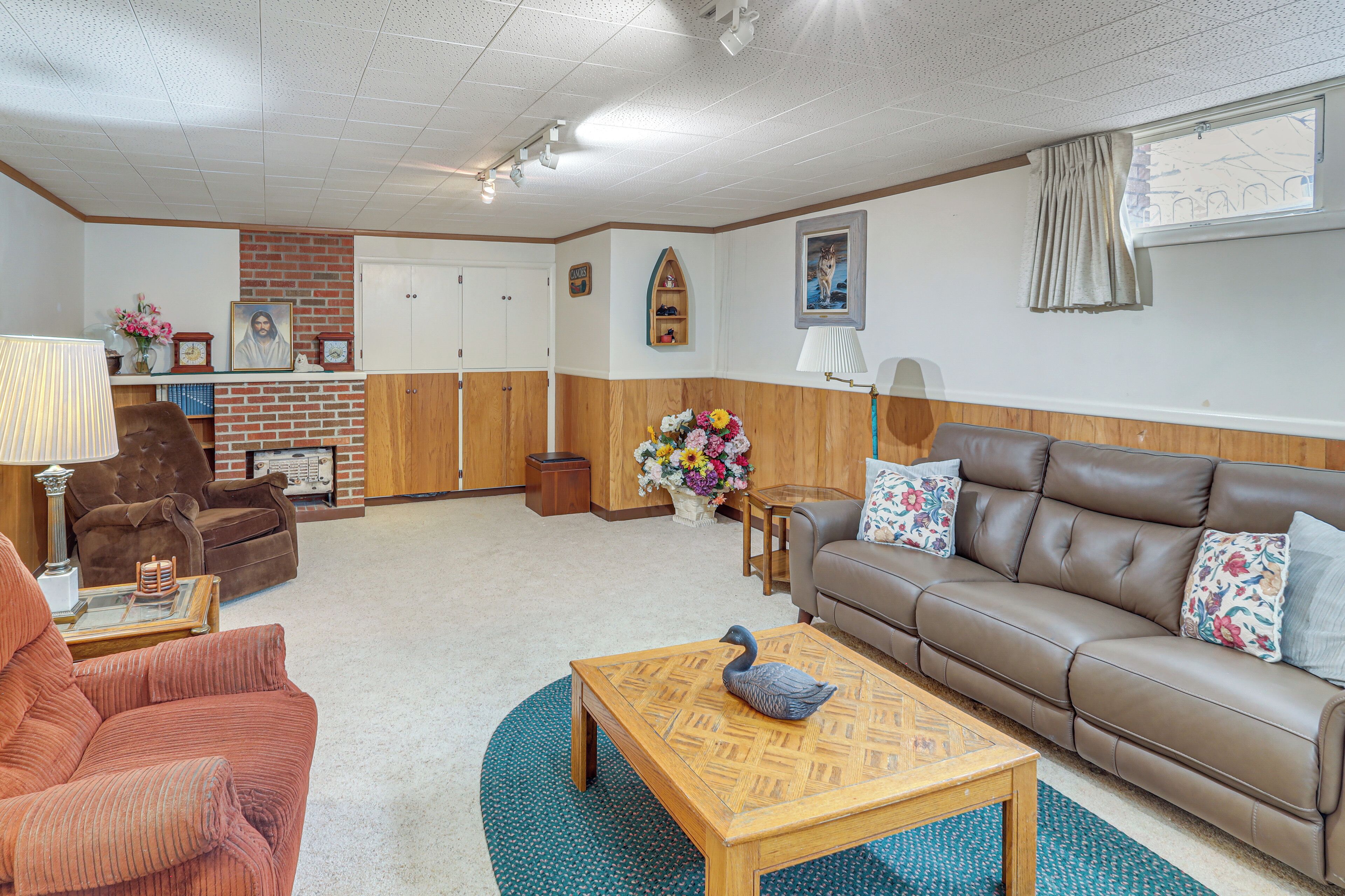 A living room with a brown leather sofa, a brown armchair, a wooden coffee table with a swan decoration, a blue patterned rug, and a brick accent wall.