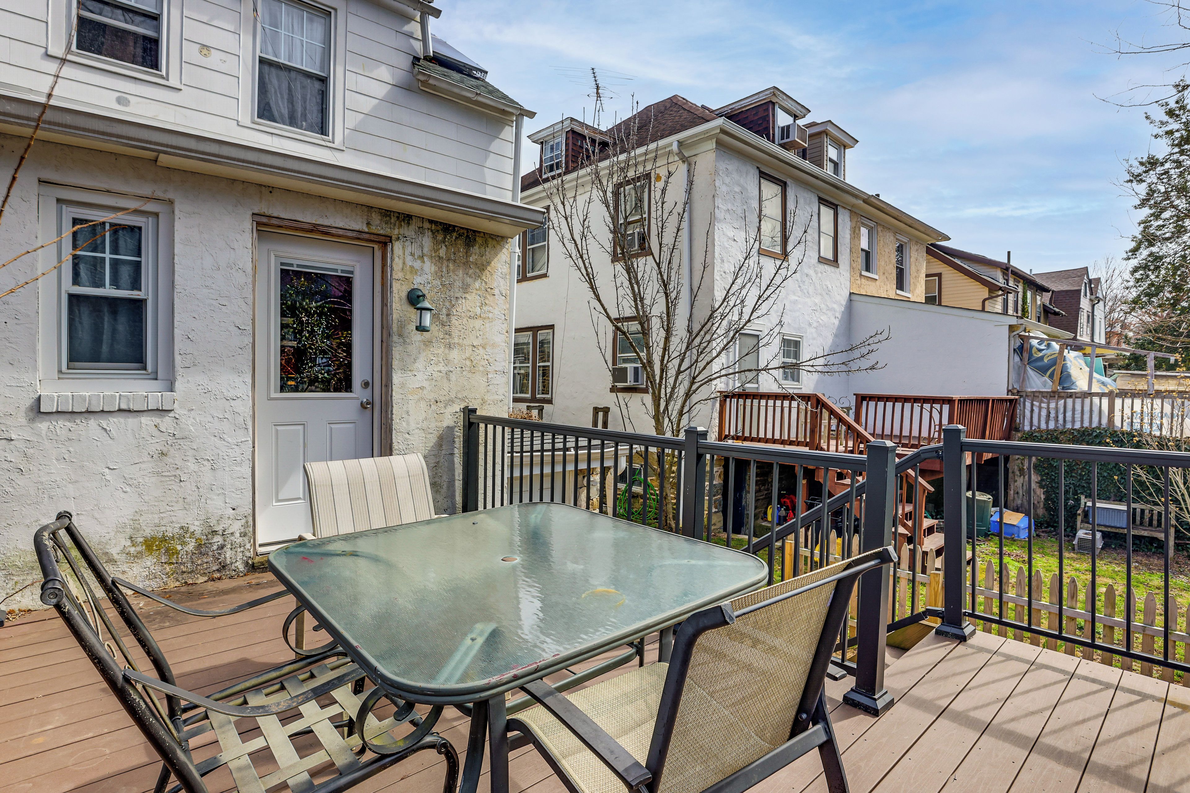 A residential deck with a glass table and chairs, a white door, and a view of neighboring houses.