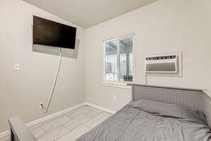 Un dormitorio con cama, televisor montado en la pared, ventana con persianas y un calentador también fijado a la pared.
