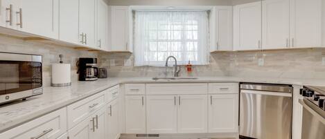 A modern kitchen with white cabinets, a stainless steel dishwasher, a built-in microwave, and a window with sheer curtains.