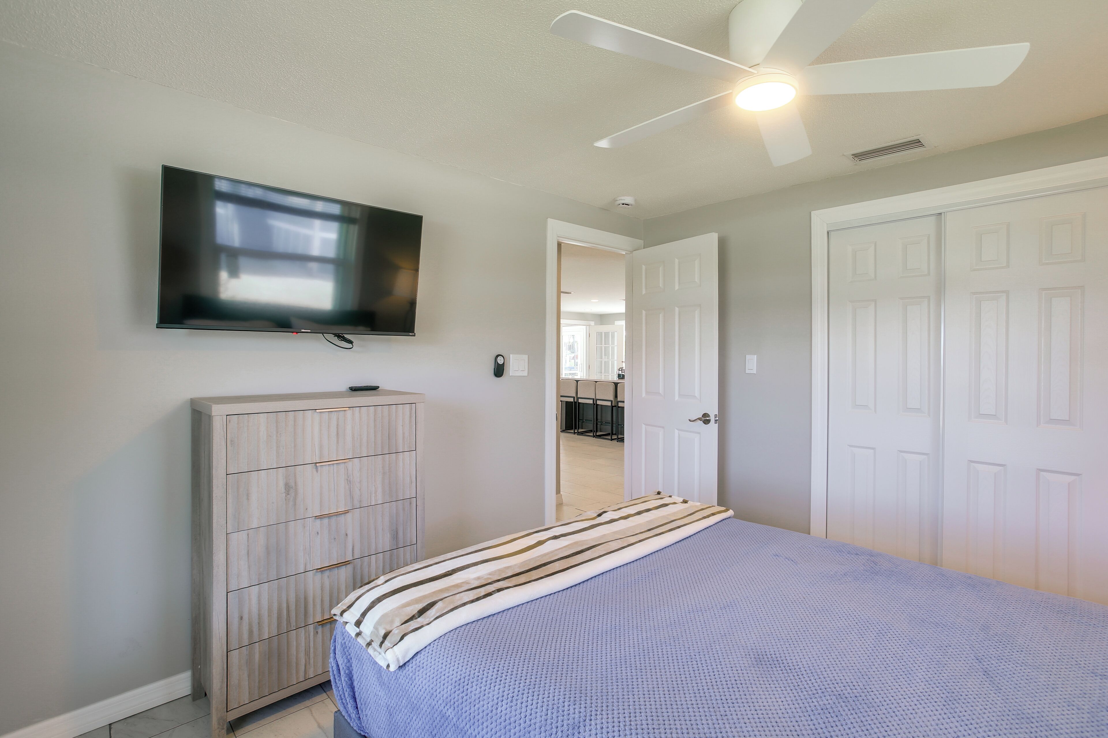 Un dormitorio con cama, televisor montado en la pared, ventilador de techo y puerta que da a otra habitación.
