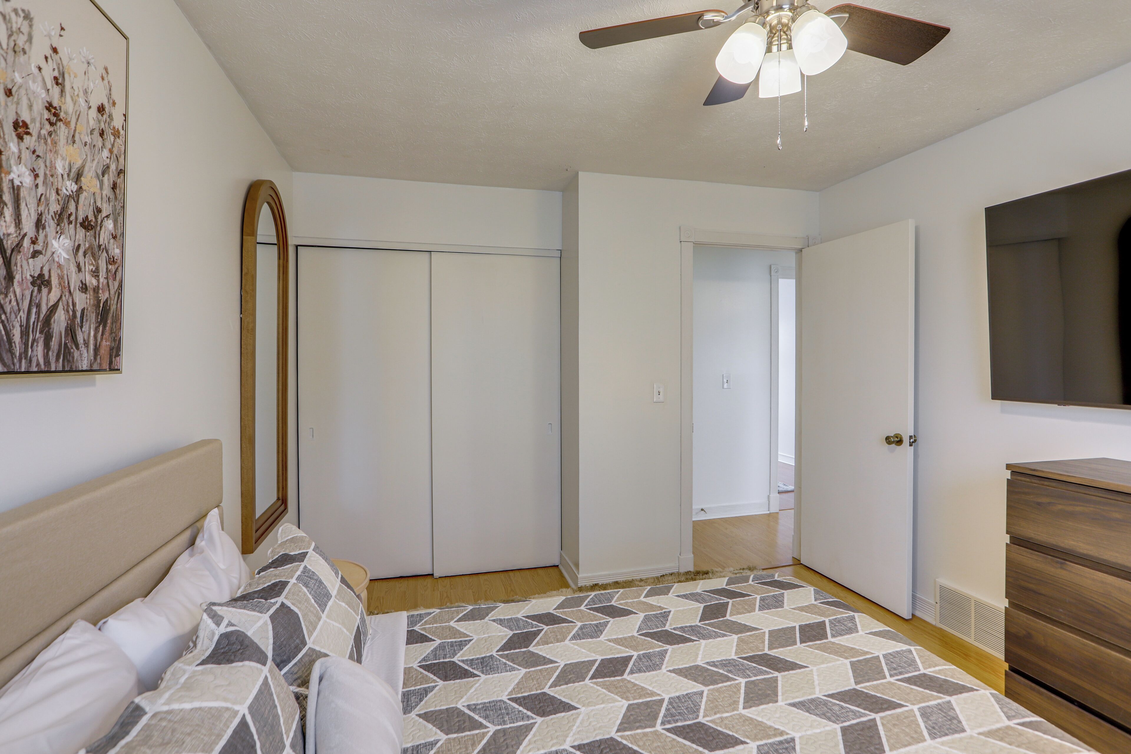 A bedroom with a bed, a dresser, a ceiling fan, and a door leading to another room.