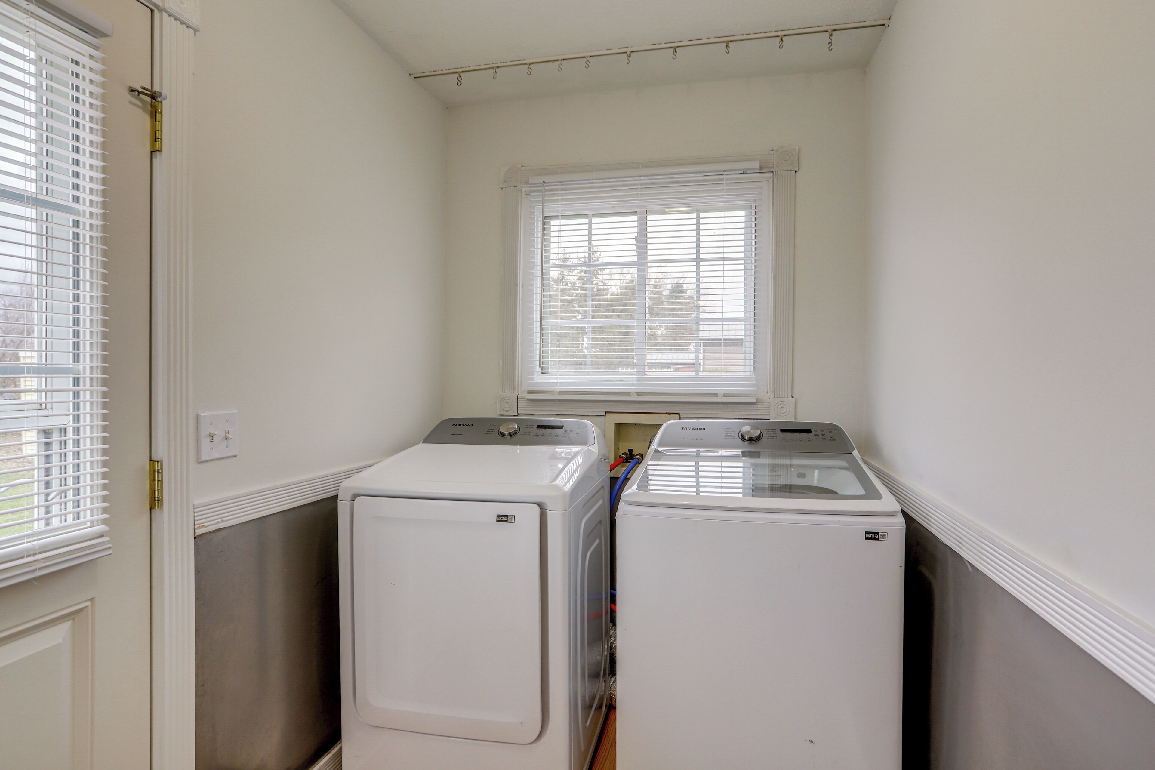 A laundry room with two front-loading washers and dryers, a window with blinds, and a door with a lock.