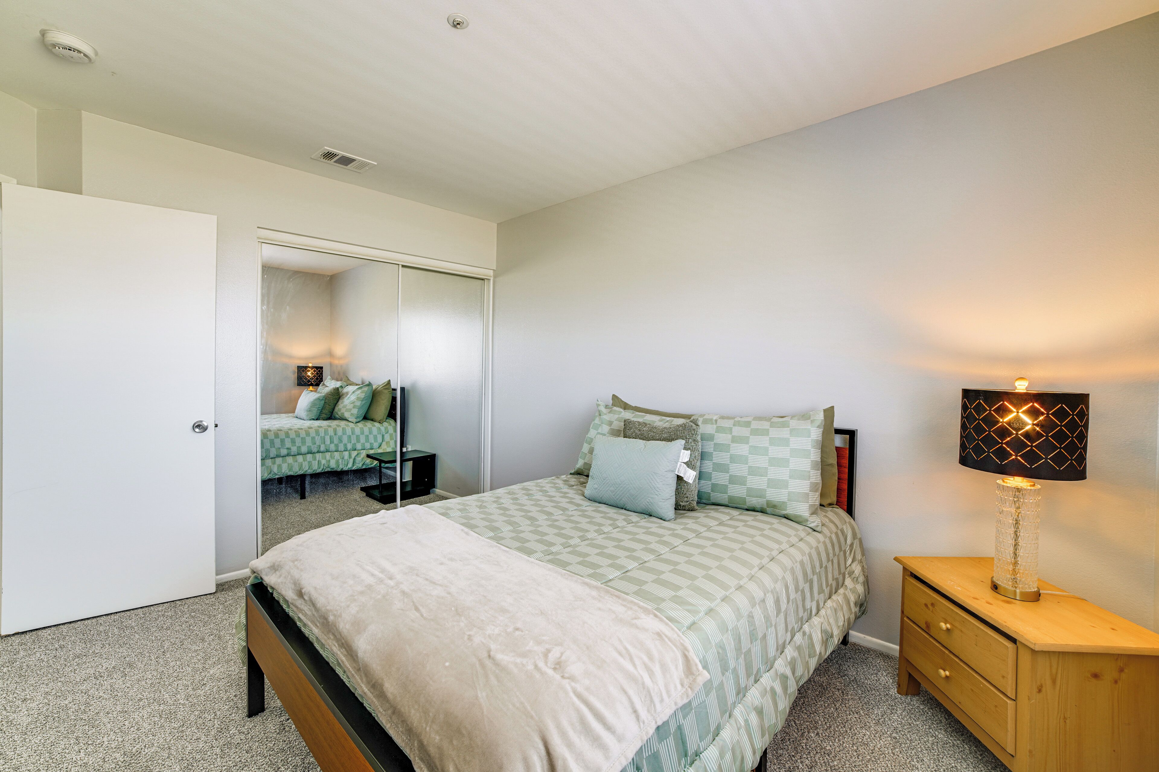 A bedroom with a bed, bedside table, lamp, and a mirror.