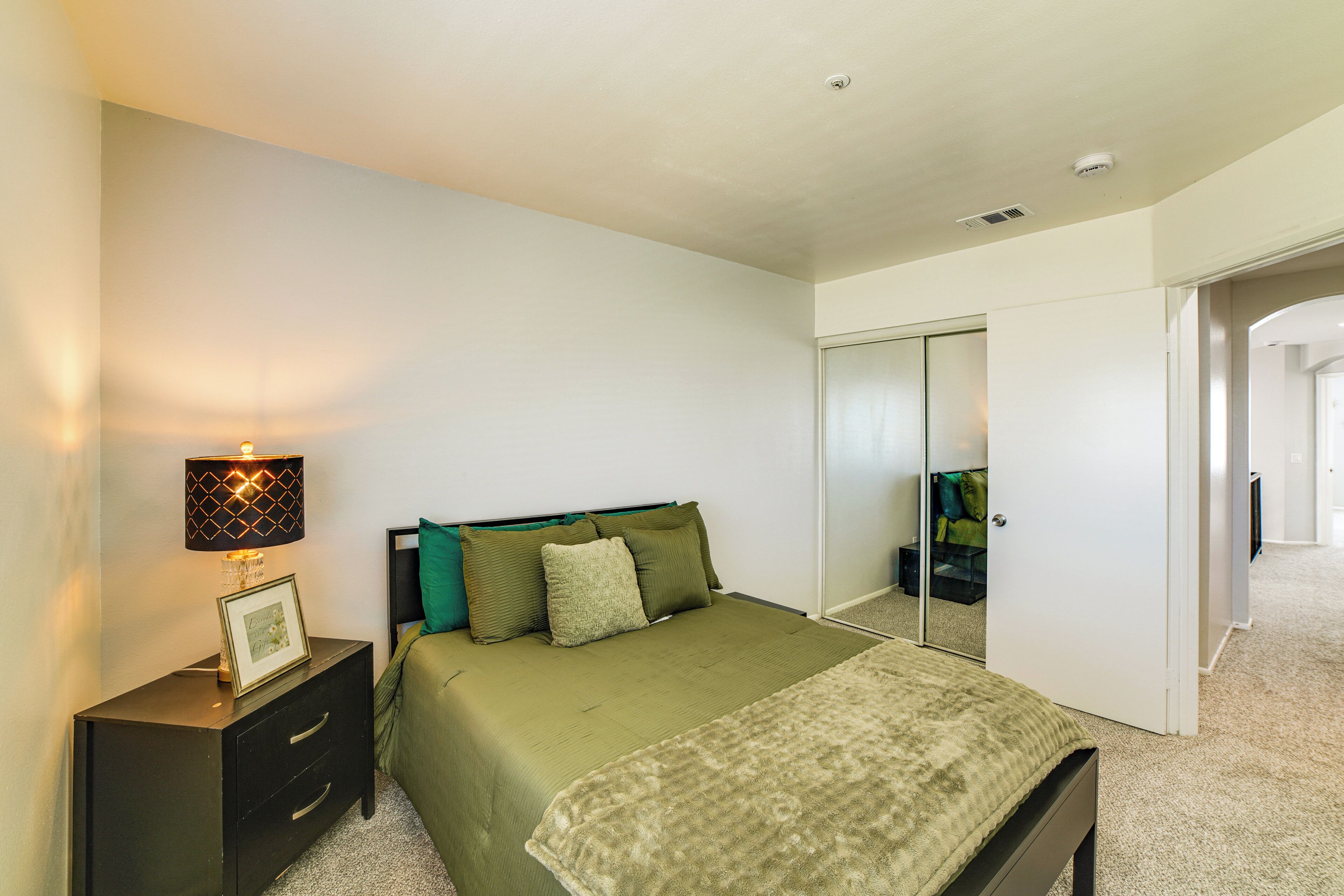 A bedroom with a bed, a bedside table with a lamp, a mirror, and a door leading to another room.