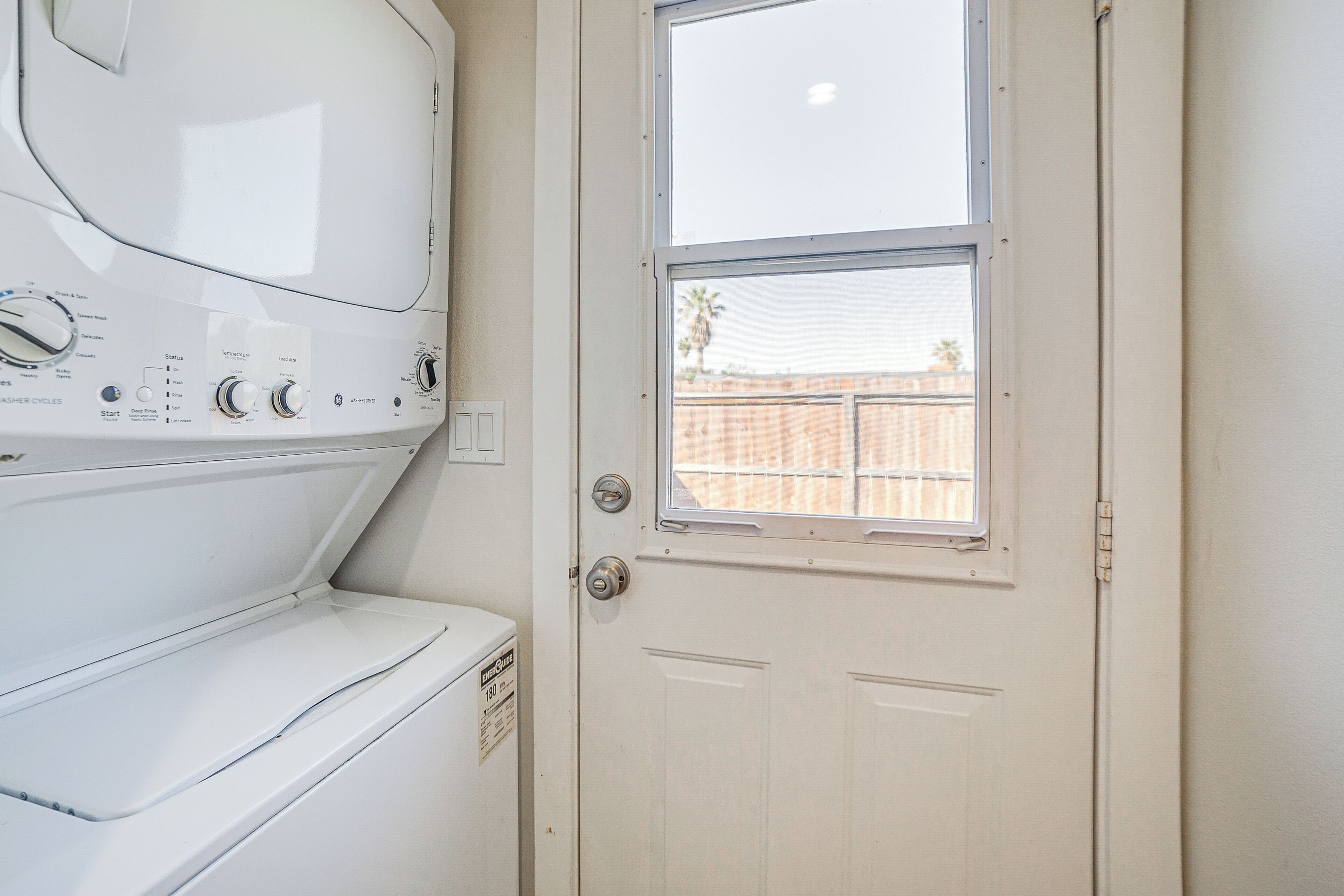 A laundry room with a front-loading washer and dryer, a door with a window, and a view of a wooden fence outside.