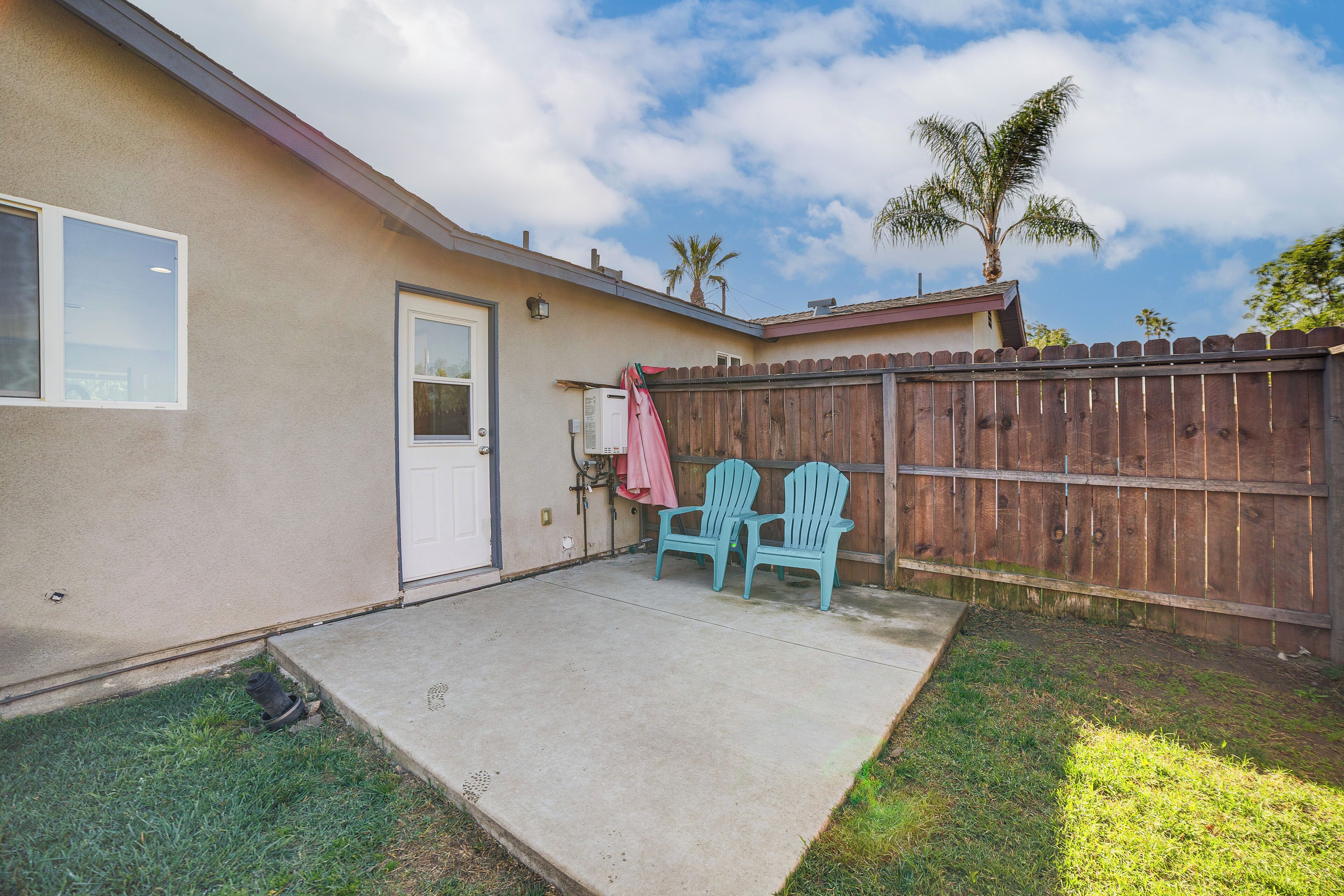 A residential backyard with a concrete patio, two Adirondack chairs, a wooden fence, and a palm tree.