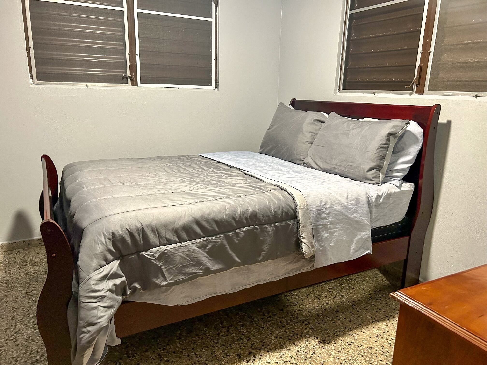 Uma cama de solteiro com estrutura de madeira e um edredom cinza, duas janelas com persianas e uma mesinha de cabeceira de madeira.