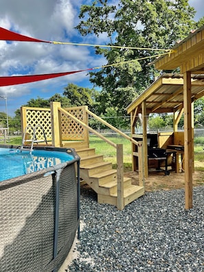 Eine Holzterrasse mit Pool, einer überdachten Grillgelegenheit und einem Spalierzaun.