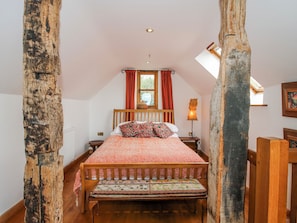 A bedroom with a sloped ceiling, a wooden bed, a bench, a nightstand, a lamp, and a window with curtains. - Bell House Cottage (Craven Arms)