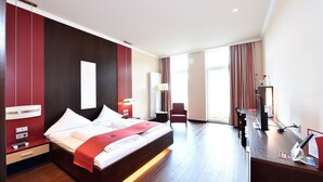 A hotel room with a large bed, a desk with a computer, a chair, and a window with curtains. - Double room Seeblick Deluxe (Heimbach)