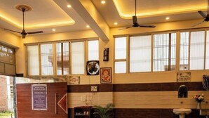 A hotel lobby with a reception desk, a bench, a potted plant, a wall-mounted clock, and a ceiling fan. - Shri Ved Vrinda Dham Aashram (Vrindavan)