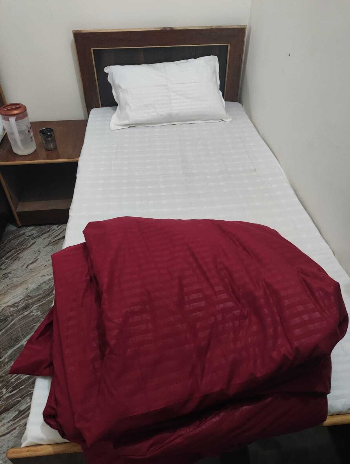 A single bed with a white pillow and a red blanket, a wooden nightstand with a bottle and a container, and a marble-patterned floor.