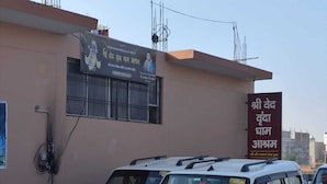 A building with a sign in a non-English script, parked white SUVs, and a brick wall. - Shri Ved Vrinda Dham Aashram (Vrindavan)