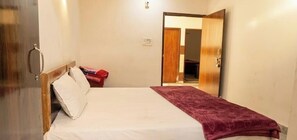 A single bed with a wooden headboard, a red blanket, and a white pillow. A door is open to a hallway. - Shri Ved Vrinda Dham Aashram (Vrindavan)