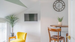 A room with a television mounted on the wall, a round wooden table with chairs, a potted plant, and a yellow chair.