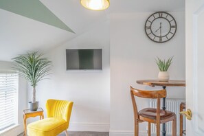 A room with a television mounted on the wall, a round wooden table with chairs, a potted plant, and a yellow chair.