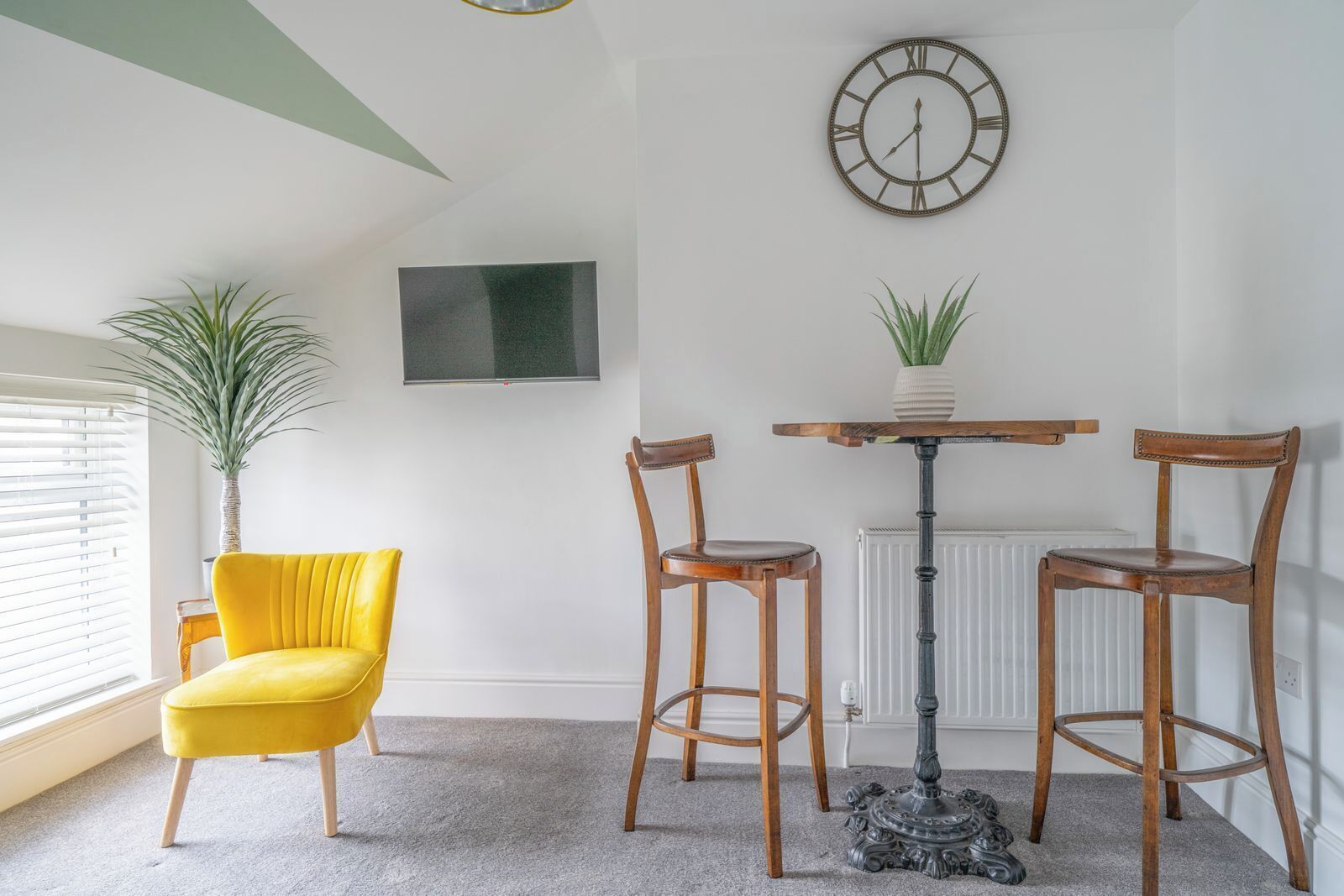 Une pièce comprenant une chaise jaune, deux chaises en bois, une table, une plante en pot, une horloge murale et un téléviseur à écran plat.