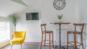 Une pièce comprenant une chaise jaune, deux chaises en bois, une table, une plante en pot, une horloge murale et un téléviseur à écran plat.