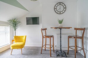 Une pièce comprenant une chaise jaune, deux chaises en bois, une table, une plante en pot, une horloge murale et un téléviseur à écran plat.