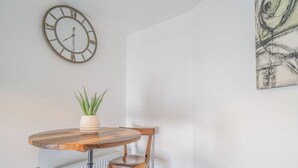 A round wooden dining table with a potted plant, two chairs, a wall clock, and a piece of abstract art.