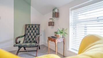 Une pièce comprenant un fauteuil en cuir capitonné, une petite table d’appoint en bois sur laquelle est posée une plante en pot, et une fenêtre avec des stores.