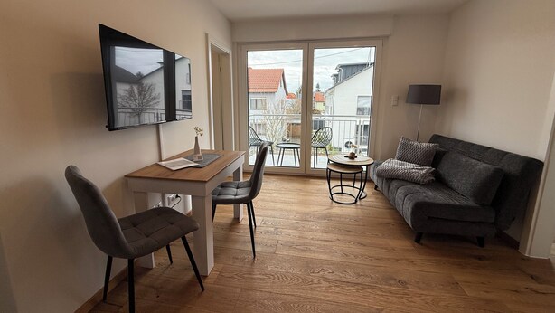 A modern living room with a sofa, a TV mounted on the wall, a dining table with chairs, and a balcony with a view of buildings. - Simon Living - Apartment Pyramide central and quiet location in Karlsruhe (Karlsruhe)