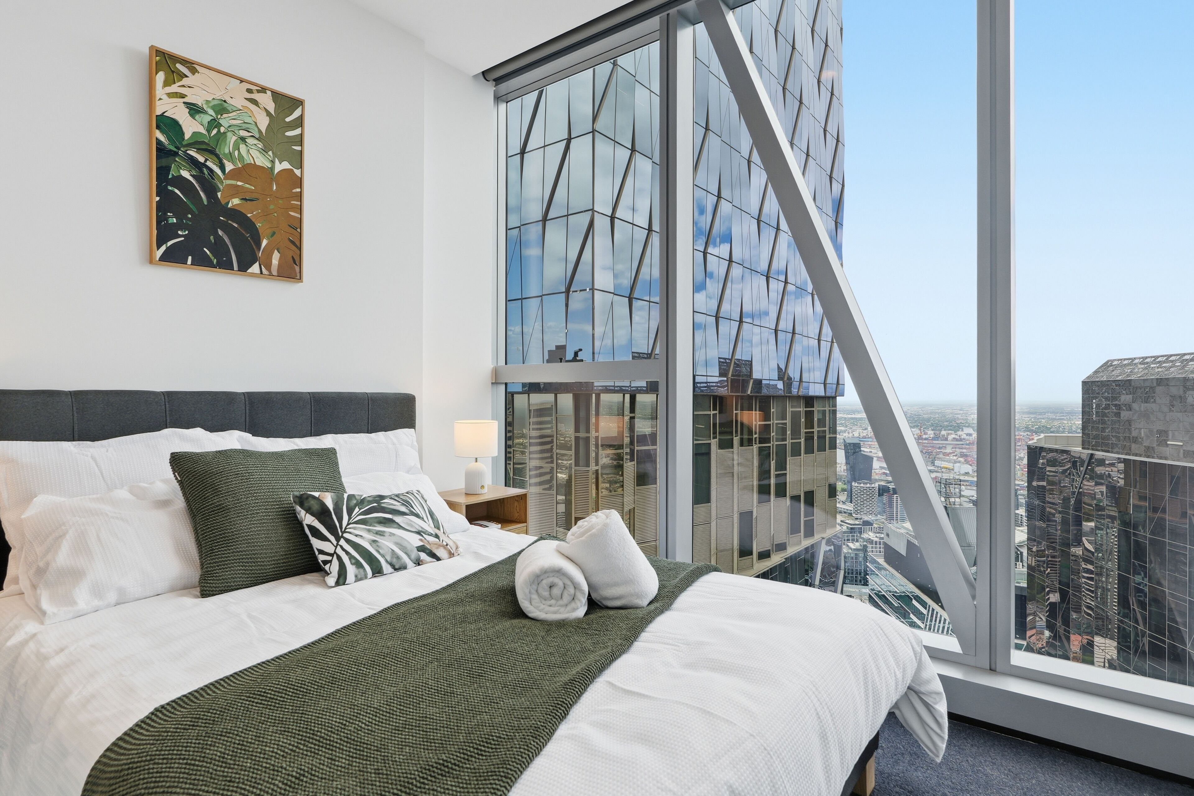 A modern hotel room with a large bed, a city view, a window with a geometric pattern, and a plant artwork on the wall.