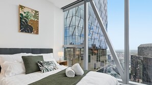 Une chambre d’hôtel moderne dotée d’un grand lit, offrant une vue sur la ville, avec une fenêtre ornée d’un motif géométrique et une œuvre d’art végétale accrochée au mur.