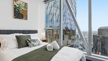 A modern hotel room with a large bed, a city view, a window with a geometric pattern, and a plant artwork on the wall.