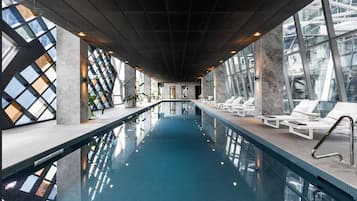 An indoor swimming pool with a long, narrow design, surrounded by lounge chairs and large windows.