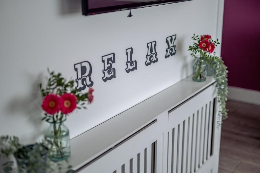 Paredes brancas com a palavra 'RELIAX' em letras estilizadas, ladeadas por dois vasos de vidro com flores vermelhas.