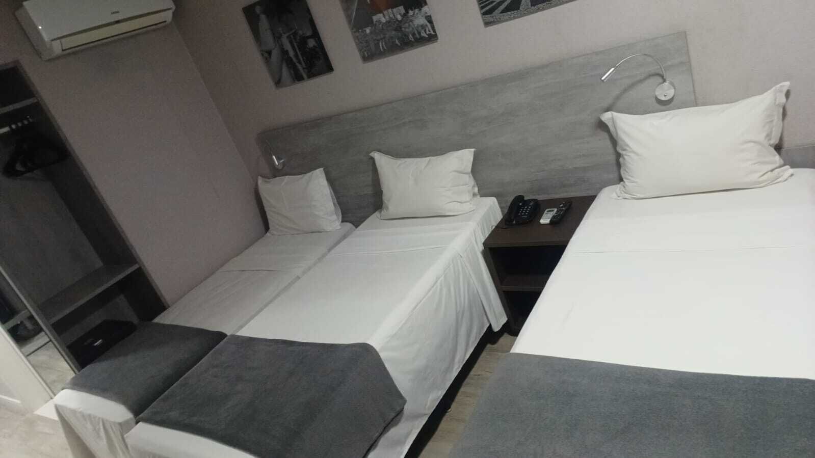 A hotel room with two single beds, a small table with a phone, a wall-mounted air conditioner, and a shelf with items.