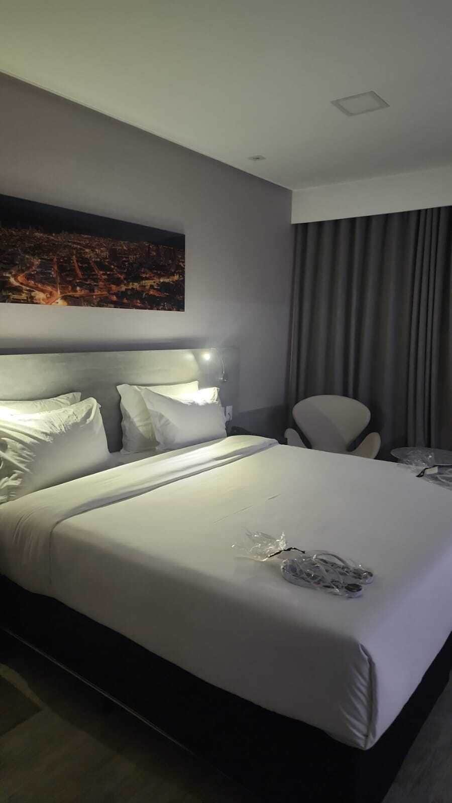 A hotel room with a large bed, a headboard, a nightstand, a chair, a painting on the wall, and a window with curtains.