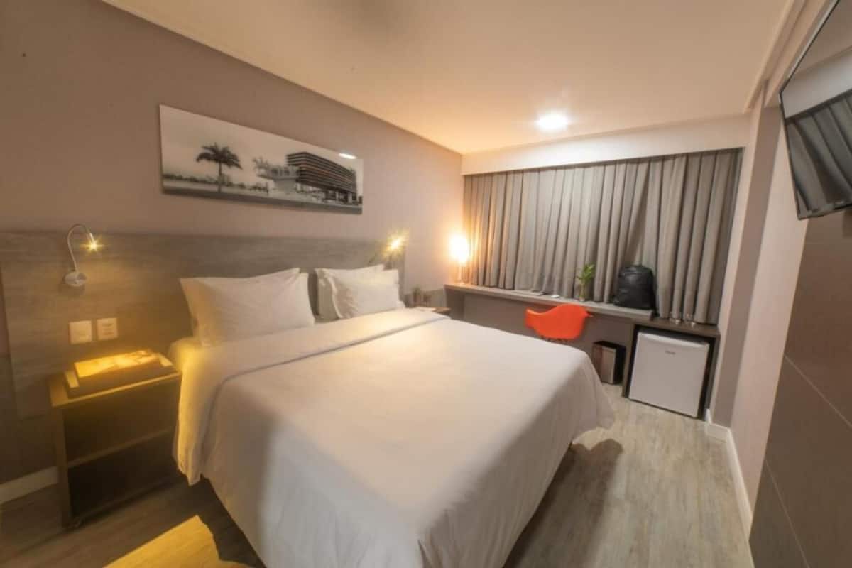 Habitación de hotel con cama, lámparas de noche, una mesita, ventana con cortinas y televisión de pared.