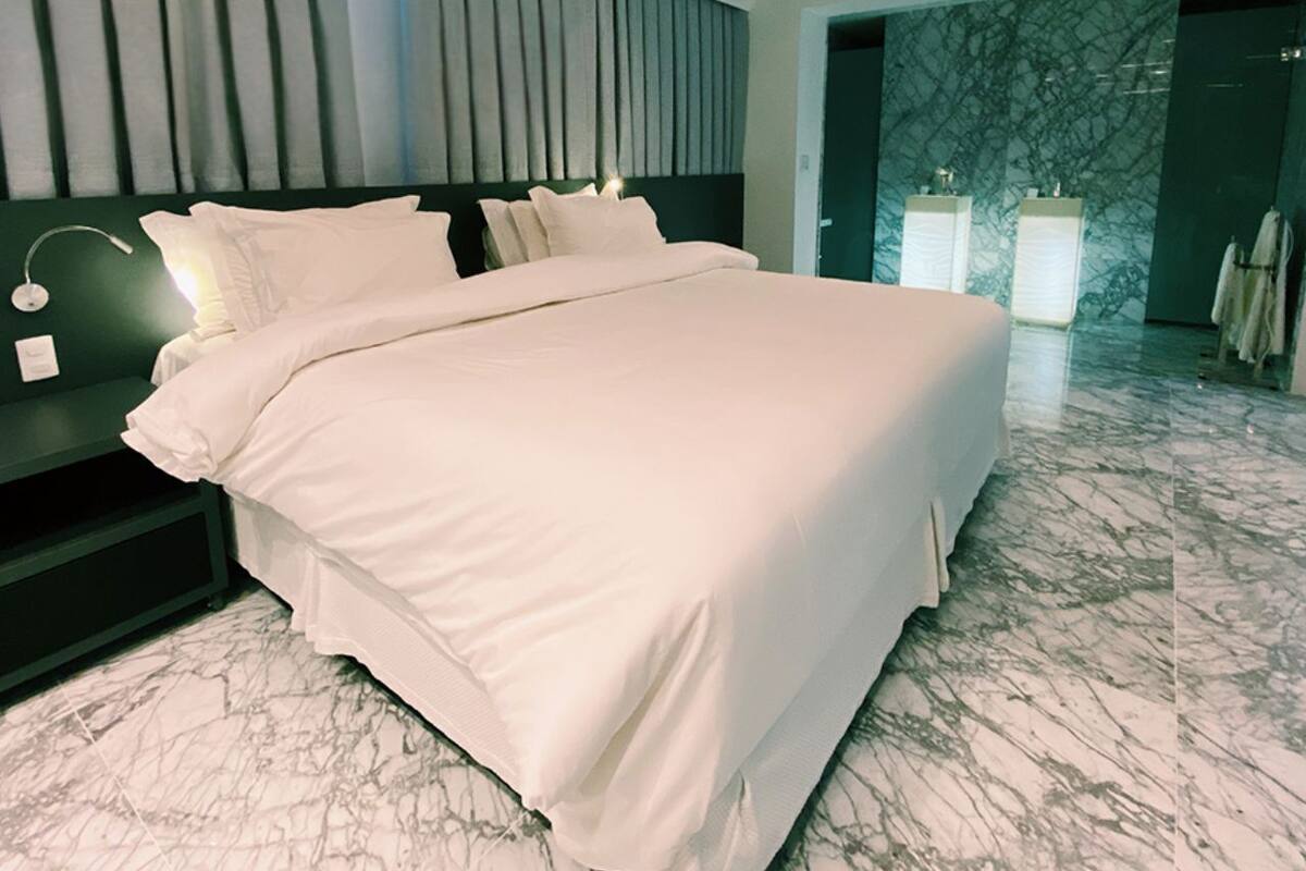 Una cama grande con sábanas blancas, un cabecero oscuro y suelo de mármol.