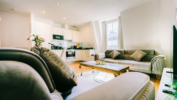 A modern living room with a sofa, coffee table, and a TV. There is a kitchen in the background with white cabinets and appliances. - LUX Bloomsbury Flat By Holborn Covent Garden (London)