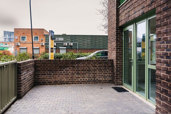 A brick building with a glass door entrance, a paved patio area, and a parked car. - LUX Riverside Duplex Masionette By LCY Excel (London)