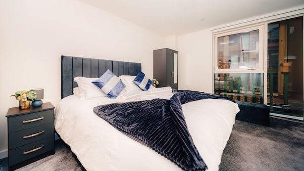 A modern bedroom with a large bed, dark headboard, and a view of a balcony with plants. - LUX Riverside Duplex Masionette By LCY Excel (London)