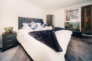 A modern bedroom with a large bed, dark headboard, and a view of a balcony with plants. - LUX Riverside Duplex Masionette By LCY Excel (London)