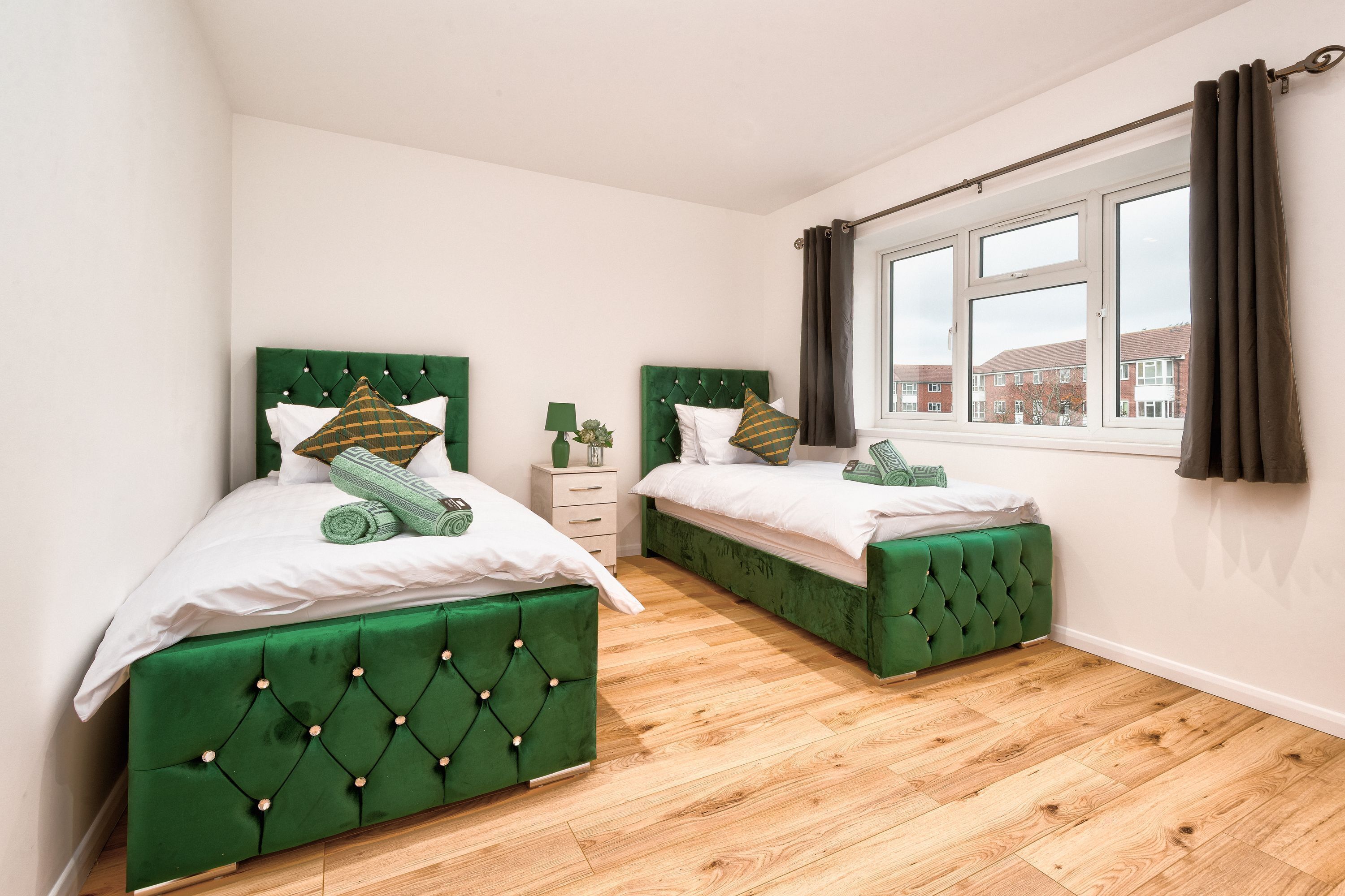 A bedroom with two green upholstered beds, a window with curtains, and a wooden floor.