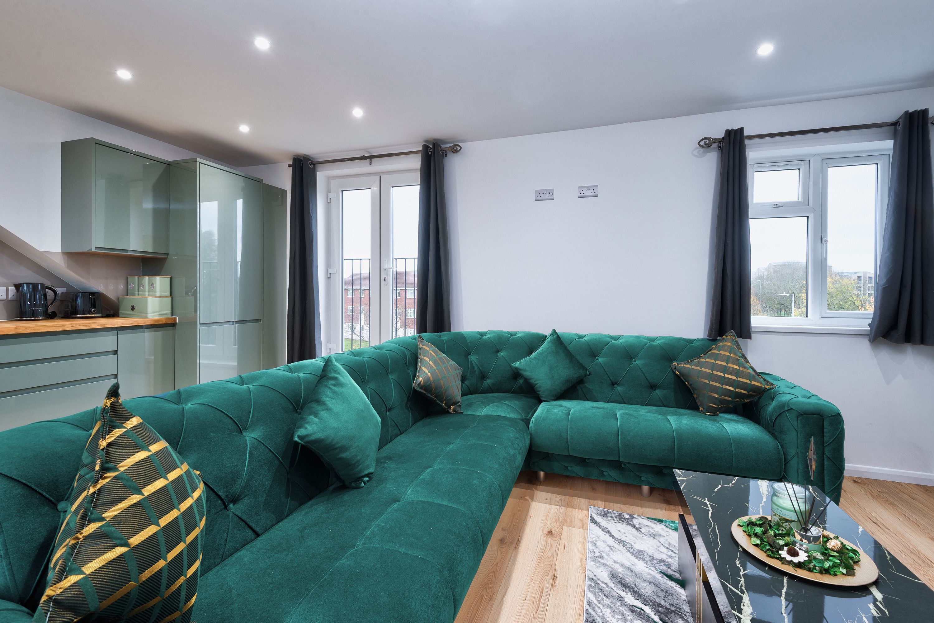 Un salon moderne avec un canapé d’angle vert, une table basse en verre et un coin cuisine doté de placards et d’un plan de travail.