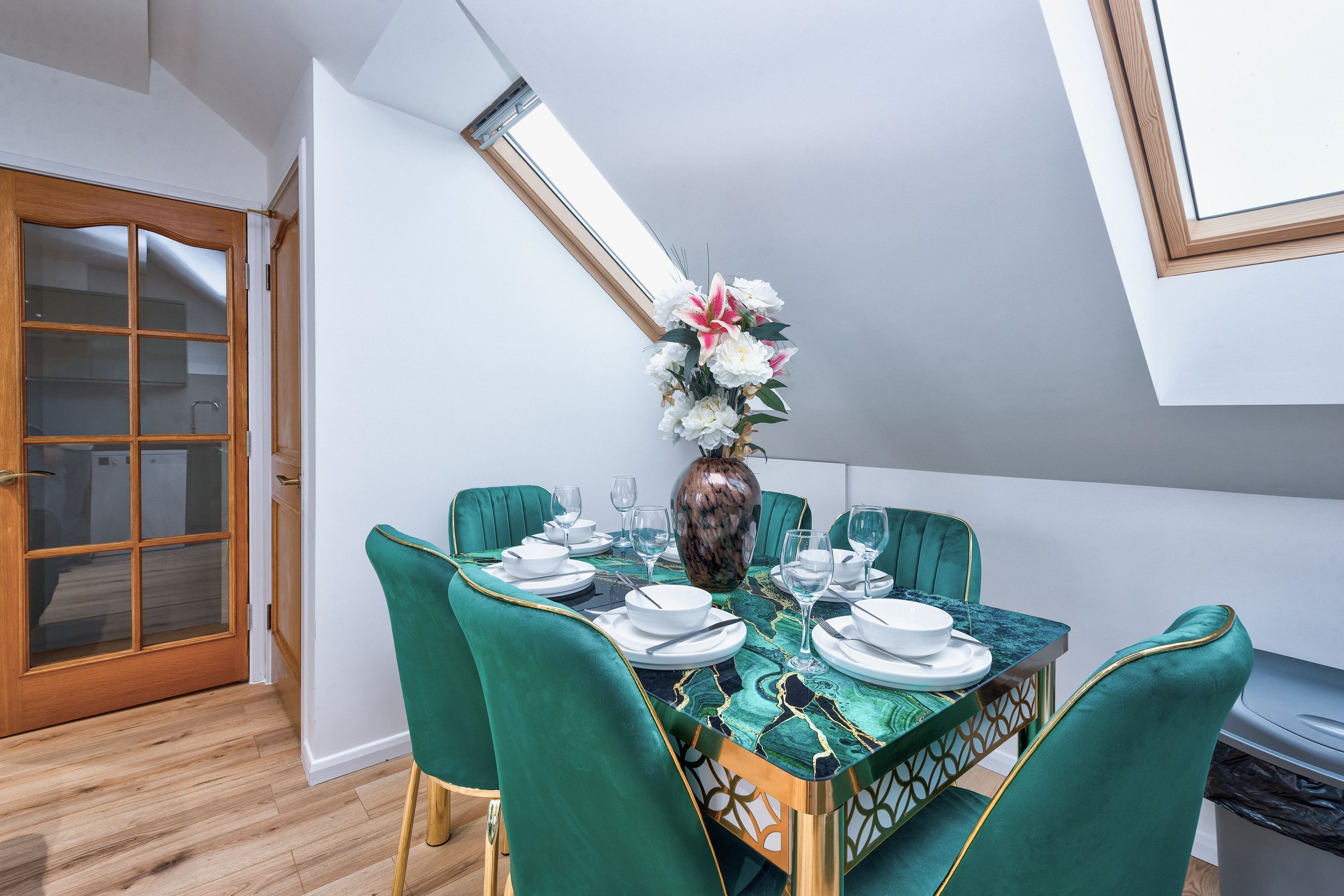 A dining area with a table set for four, green upholstered chairs, a vase with flowers, and a view of the sky through a skylight.