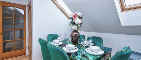 A dining area with a table set for four, green upholstered chairs, a vase with flowers, and a view of the sky through a skylight.