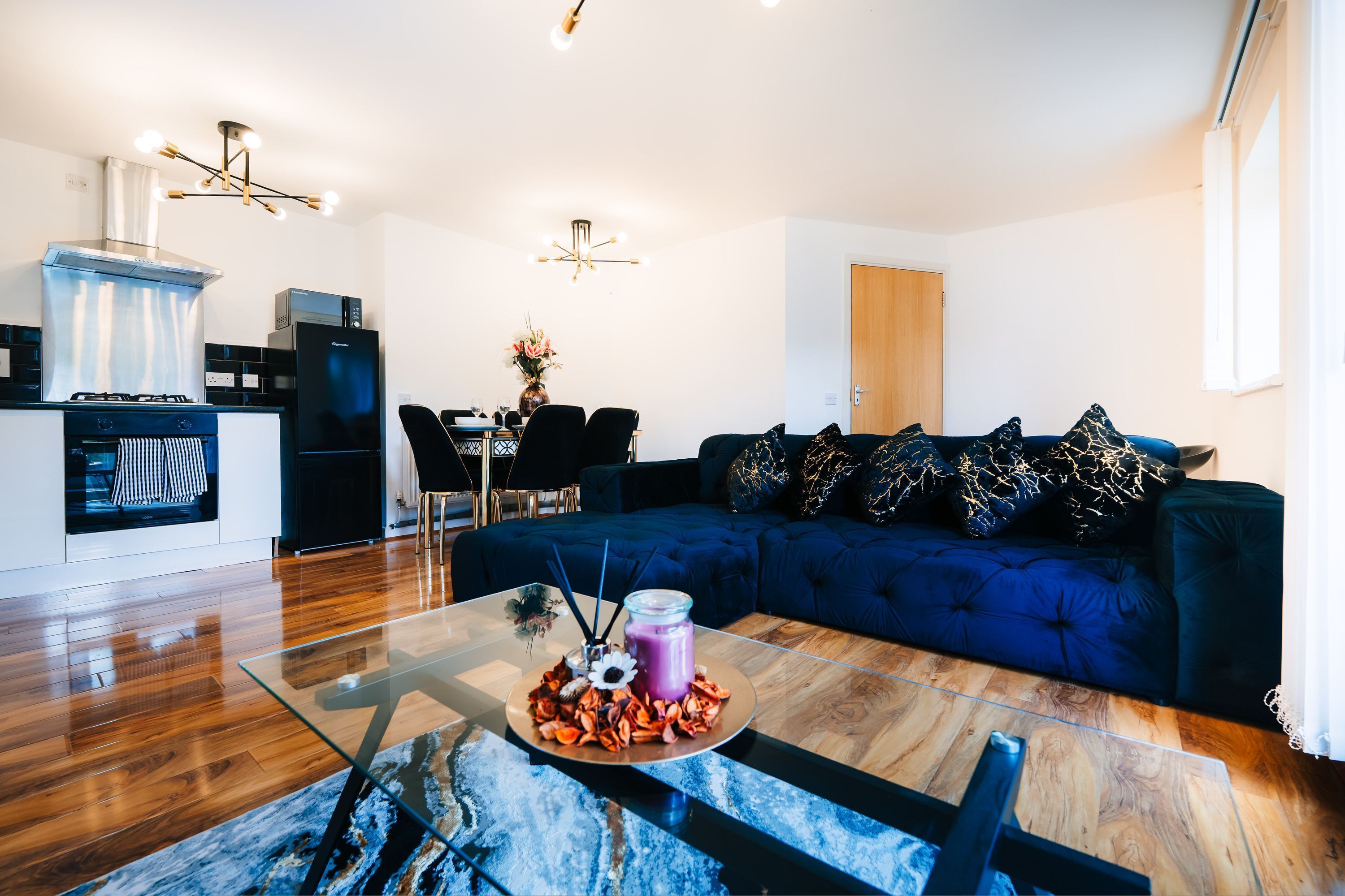 A modern living room with a glass coffee table, a blue sofa, and a dining area with chairs and a table.