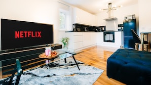 A modern living room with a flat-screen TV displaying 'NETFLIX,' a glass coffee table, a dark blue sofa, and a kitchen area with white cabinets and black appliances. - LUX Woolwich Flat 2 Stops to Canary Wharf (London)