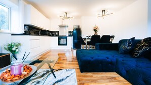 A modern kitchen with white cabinets, a black refrigerator, and a dining area with a dark blue sofa and a vase of flowers. - LUX Woolwich Flat 2 Stops to Canary Wharf (London)