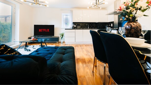 A modern living room with a sofa, a dining area with a table and chairs, a kitchen with white cabinets, a TV displaying 'NETFLIX,' and a vase with flowers. - LUX Woolwich Flat 2 Stops to Canary Wharf (London)