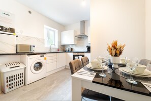 A modern kitchen with a washing machine, microwave, and dining area with a table set for two. - LUX 2bdr Flat Near LTN Airport City Centre (Luton)