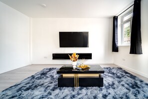 Ein modernes Wohnzimmer mit einem an der Wand befestigten Flachbildfernseher, einem schwarzen Couchtisch mit einer Vase und Kerzen sowie einem flauschigen blauen Teppich.